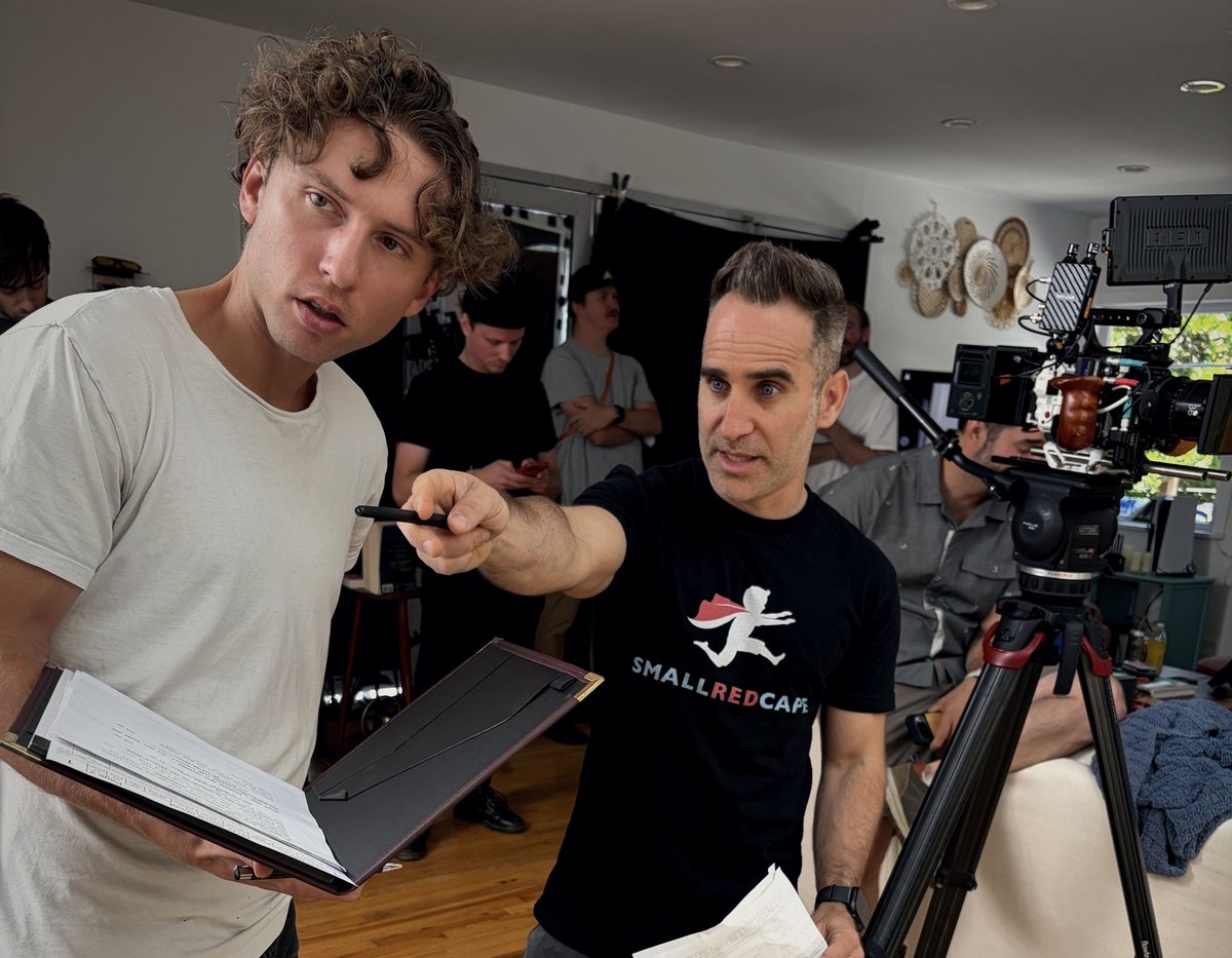 Ben in a Small Red Cape shirt on set, pointing as he directs an actor holding a script.