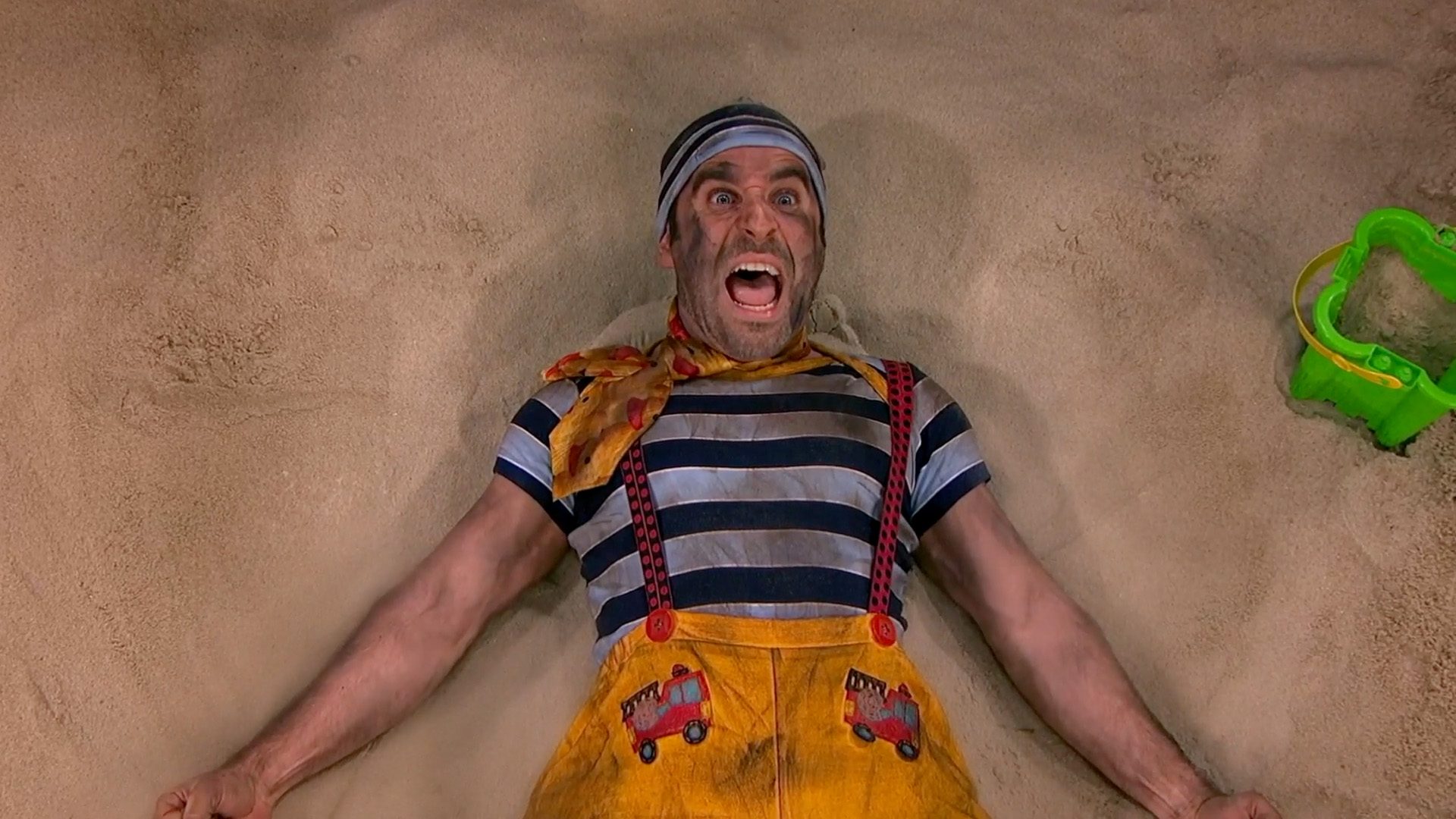 Ben as The Toddler, sprawled in a sandbox in a striped shirt and overalls, mid-scream.